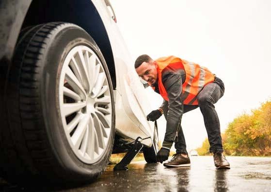 Man in an orange safety vest changing a car tire on a roadside with a jack.