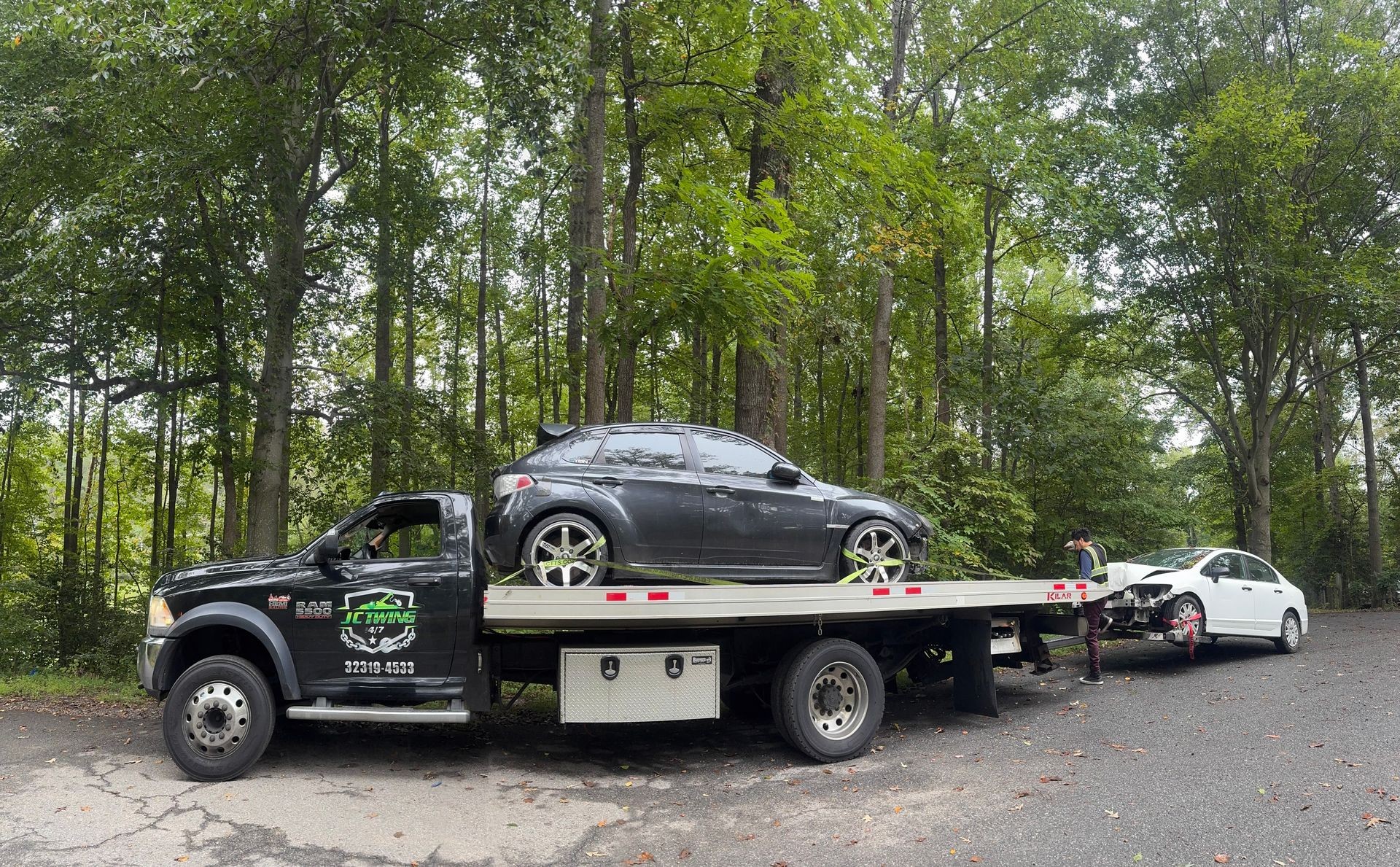 Tow truck transporting two cars through a forested area on a road.