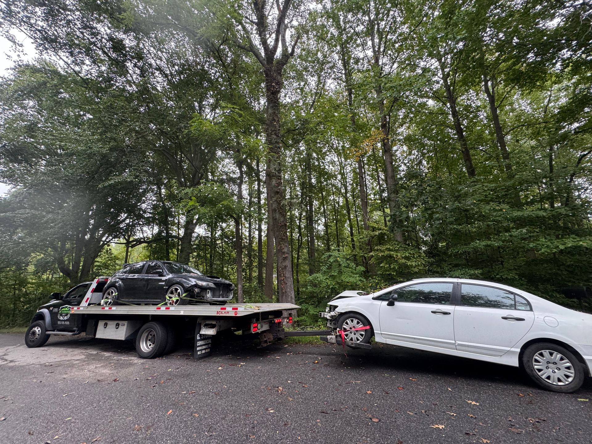 Tow truck carrying a damaged black car with another car hitched behind, surrounded by trees.