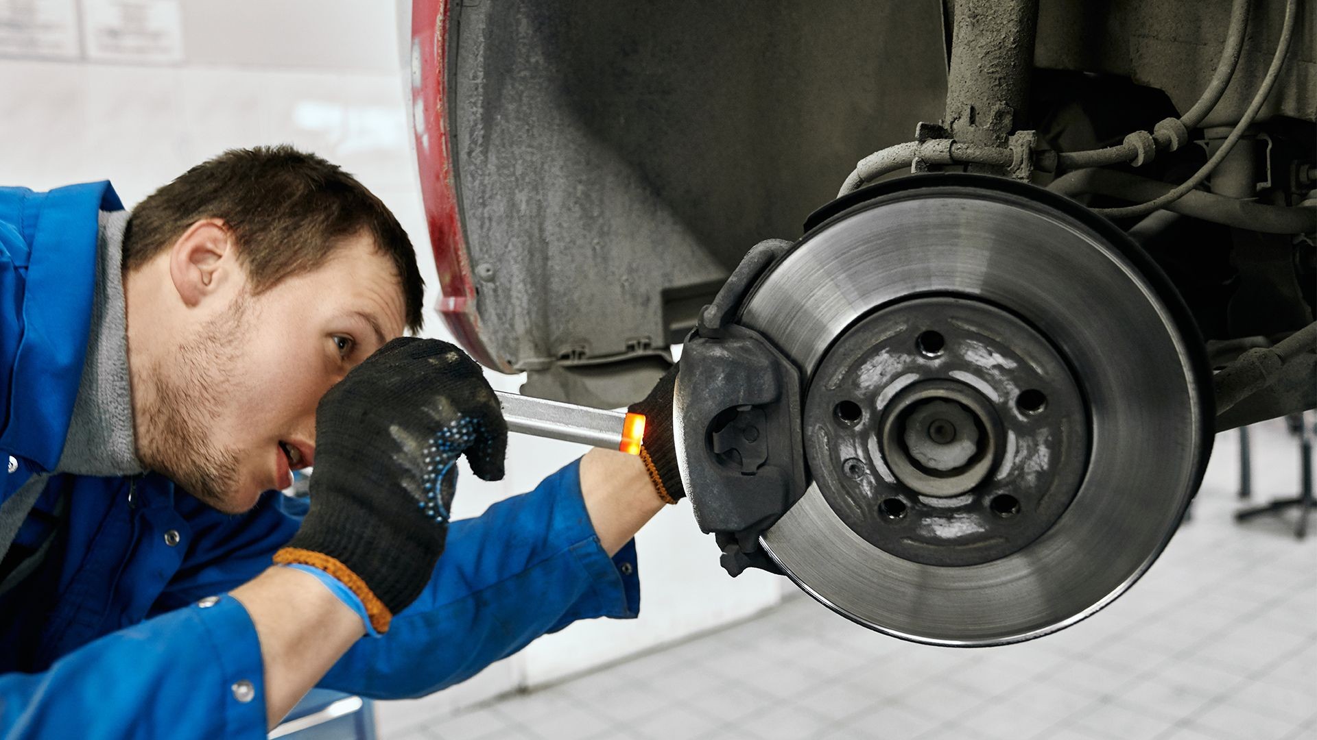 Mechanic in blue uniform inspecting a car's brake disc and caliper with a flashlight.