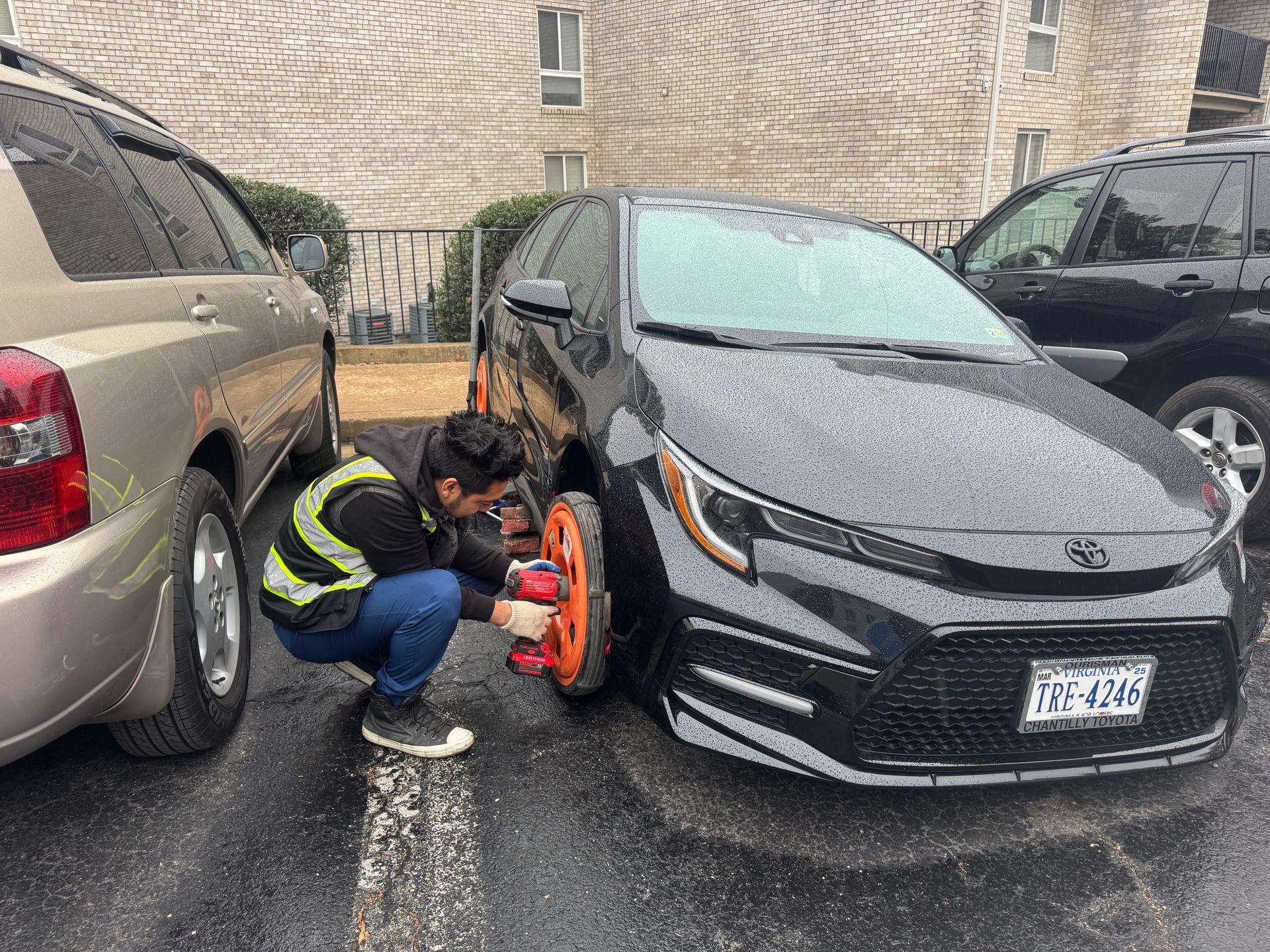 Person fixing a flat tire on a black car using an orange spare tire in a parking lot.