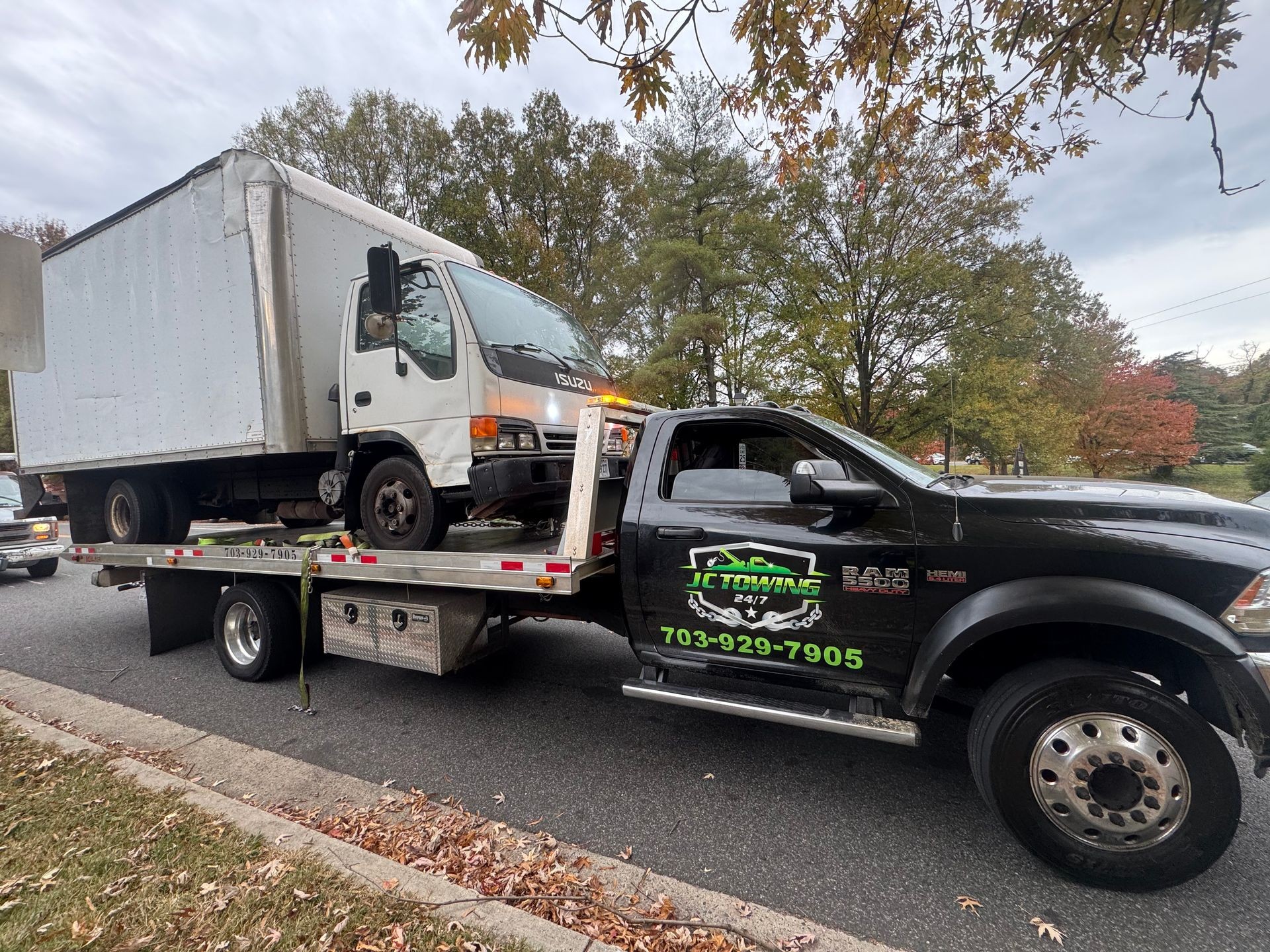 Tow truck carrying a white delivery truck on a suburban street with autumn trees in the background.